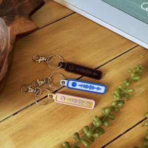 May include: Three keychains featuring Spotify code designs. One is black with a red border, another is blue with a white border, and the third is pink with a yellow border. Each keychain has a silver keyring and clasp.