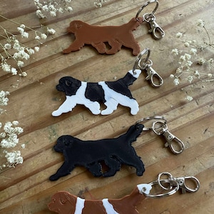 May include: Four dog-shaped keychains in various colors: brown, black and white, black, and brown and white. Each keychain has a silver-colored keyring and clasp. The keychains are arranged on a wooden surface with white flowers.