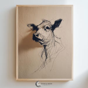 Puede incluir: Una obra de arte enmarcada que presenta un boceto de la cabeza y los hombros de una vaca. La vaca está representada con líneas negras sobre un fondo beige. La obra de arte está en un marco de madera de color claro. La parte inferior del marco tiene el texto "STONE & MOON PRINTS".