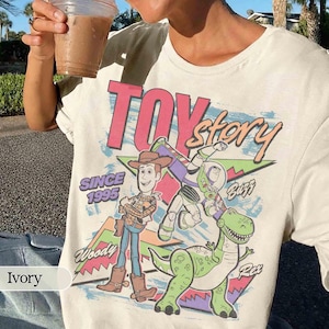 May include: Ivory-colored t-shirt featuring a graphic of characters from the animated film "Toy Story." The design includes Woody, Buzz Lightyear, and Rex, with the text "TOY Story" and "SINCE 1995."