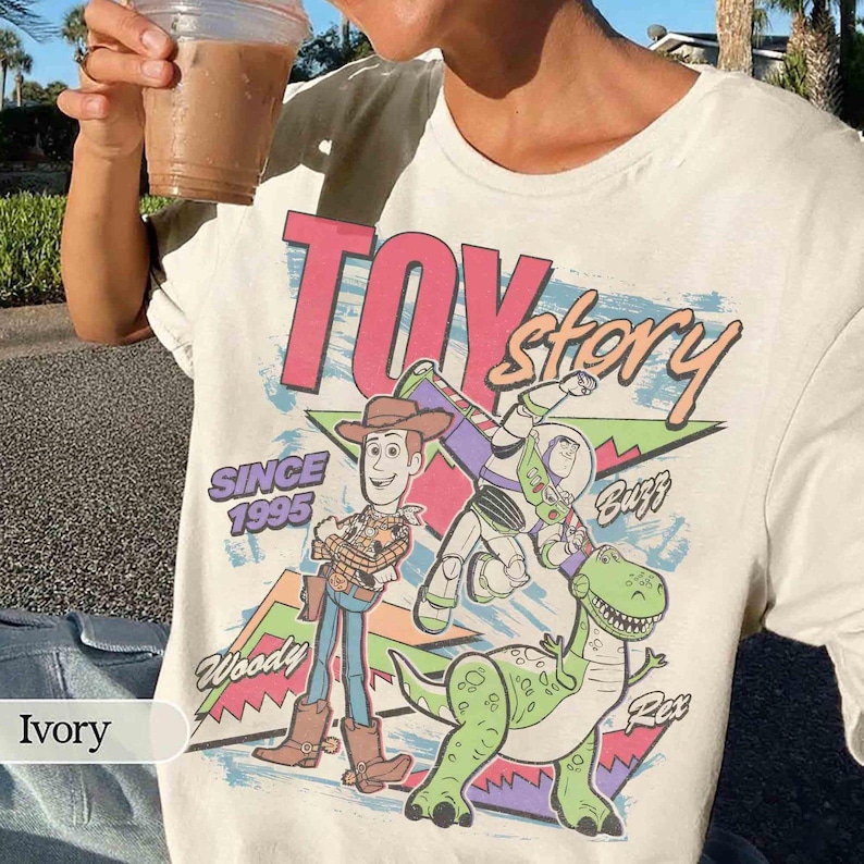May include: Ivory-colored t-shirt featuring a graphic of characters from the animated film "Toy Story." The design includes Woody, Buzz Lightyear, and Rex, with the text "TOY Story" and "SINCE 1995."