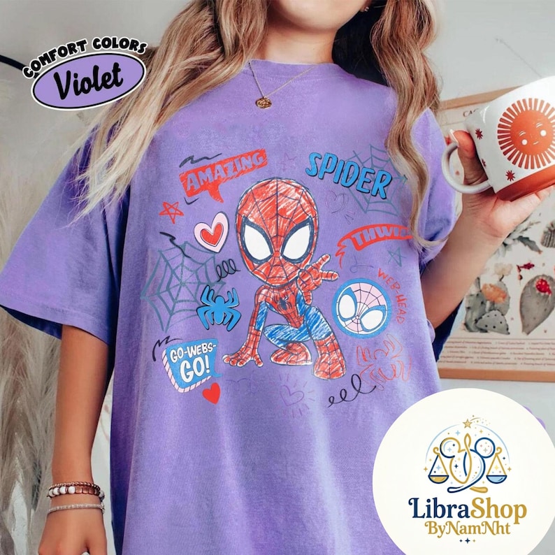 Puede incluir: Camiseta violeta Comfort Colors con un gr&aacute;fico de Spider-Man. El dise&ntilde;o incluye la palabra "AMAZING" en un bocadillo, un coraz&oacute;n, telara&ntilde;as y las palabras "GO-WEBS-GO!" y "SPIDER". La camiseta tambi&eacute;n tiene el logo de LibraShop.