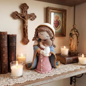 Puede incluir: Muñeca de ganchillo hecha a mano de la Virgen María sosteniendo al niño Jesús, con un vestido rosa y un chal azul. La muñeca está en un estante con velas, libros y arte religioso. Un crucifijo de madera cuelga de la pared.