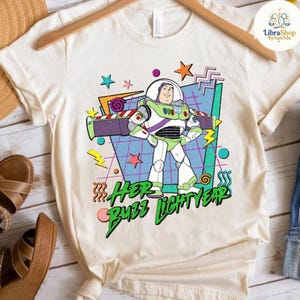 May include: Cream-colored t-shirt featuring a graphic of Buzz Lightyear from Toy Story. The design includes the text "Her Buzz Lightyear" in green and pink, with stars, lightning bolts, and geometric shapes in the background.
