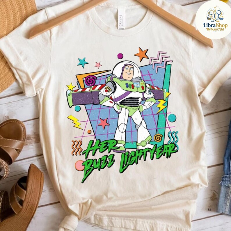 May include: Cream-colored t-shirt featuring a graphic of Buzz Lightyear from Toy Story. The design includes the text "Her Buzz Lightyear" in green and pink, with stars, lightning bolts, and geometric shapes in the background.