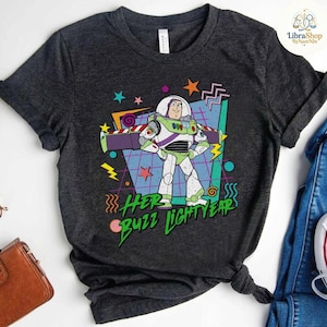 May include: Dark gray t-shirt featuring a colorful graphic of Buzz Lightyear from Toy Story. The design includes the text "Her Buzz Lightyears" in green, with stars, lightning bolts, and geometric shapes. The shirt is made of a soft material.