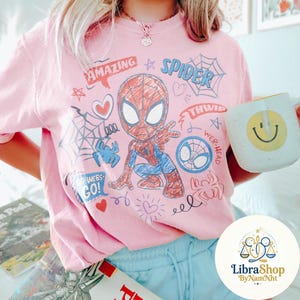Puede incluir: Sudadera rosa claro con un dise&ntilde;o colorido de Spider-Man, con las palabras "AMAZING" y "SPIDER". El dise&ntilde;o incluye telara&ntilde;as, corazones y las frases "GO-WEBS-GO!" y "WEB-HEAD". Tambi&eacute;n se ve una taza blanca con una carita sonriente amarilla.