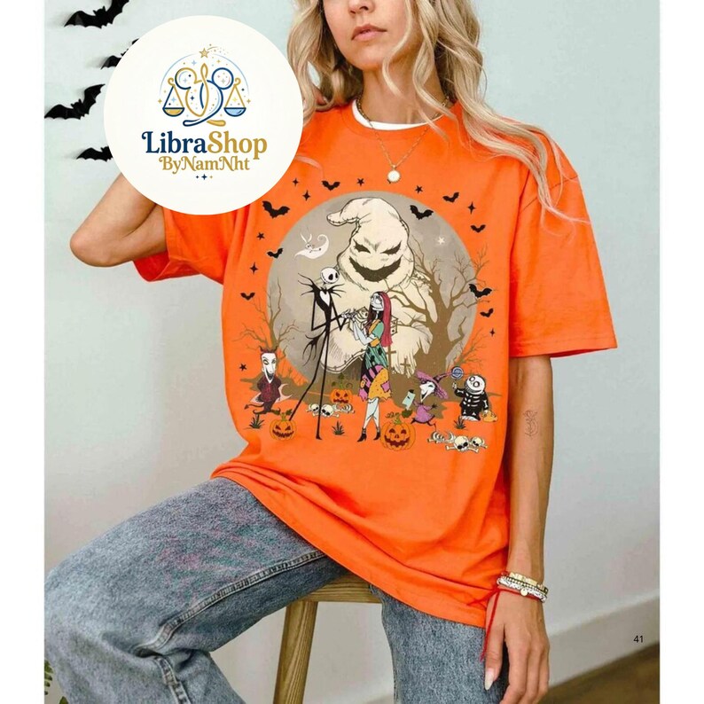 Może przedstawiać: Pomarańczowa koszulka z grafiką z filmu "Miasteczko Halloween". Projekt zawiera Jacka Skellingtona, Sally, Oogie Boogie i inne postacie. Koszulka ma okrągły dekolt i kr&oacute;tkie rękawy. Logo LibraShop jest widoczne w lewym g&oacute;rnym rogu.