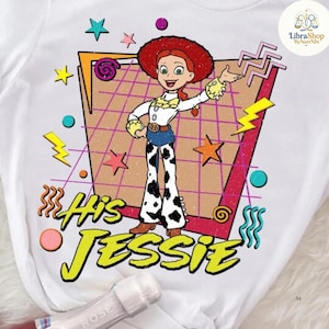 May include: White t-shirt featuring a graphic of Jessie from Toy Story. The design includes a retro-style background with stars, lightning bolts, and the text "His Jessie" in yellow. Jessie is wearing a red hat, white shirt, blue jeans, and cow-print pants.