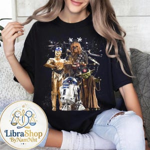 Może przedstawiać: Czarny t-shirt z grafiką Star Wars przedstawiającą C-3PO, Chewbaccę, R2-D2 i Yodę. Projekt zawiera gwiezdne tło i świąteczne elementy. Wygodny, codzienny element garderoby.
