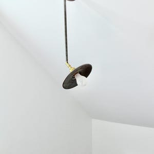 May include: A vintage-style pendant light fixture. It features a circular, dark metal shade with a visible lightbulb. The fixture is suspended from the ceiling by a slender, dark metal rod and a circular ceiling mount.