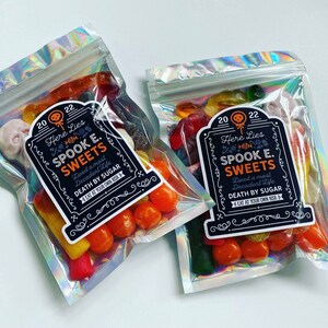 May include: Two cellophane bags with a holographic sheen, filled with Halloween sweets. Each bag features a label reading "Here Lies Spook E. Sweets, Lived a most Decadent Life, Death by Sugar, Eat at Your Own Risk."