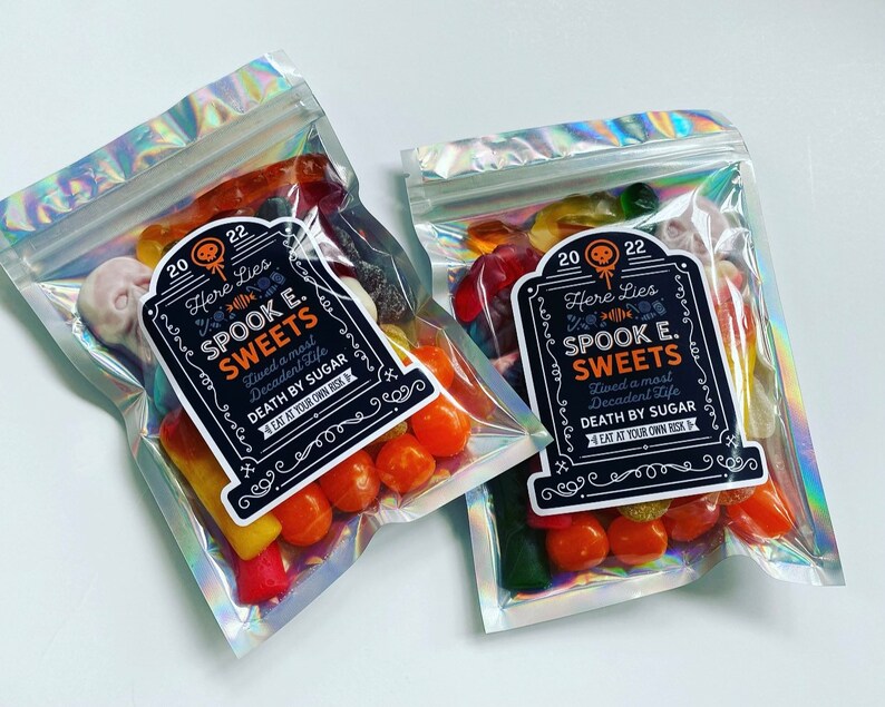 May include: Two cellophane bags with a holographic sheen, filled with Halloween sweets. Each bag features a label reading "Here Lies Spook E. Sweets, Lived a most Decadent Life, Death by Sugar, Eat at Your Own Risk."