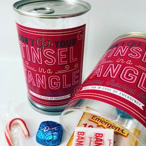 May include: Two clear plastic containers shaped like cans with red labels that read "Don't Get Your Tinsel in a Tangle." One is upright, the other tilted. Contents include candy, bandages, and Emergen-C packets.