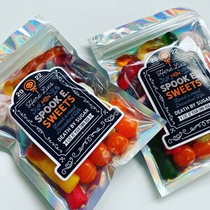 May include: Two cellophane bags filled with colourful sweets. Each bag has a black label with the text "Here Lies Spook E. Sweets" and "Death by Sugar". The sweets include orange, yellow, red, and green gummy treats, perfect for Halloween.