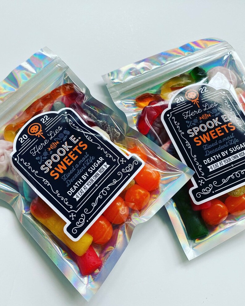 May include: Two cellophane bags filled with colourful sweets. Each bag has a black label with the text "Here Lies Spook E. Sweets" and "Death by Sugar". The sweets include orange, yellow, red, and green gummy treats, perfect for Halloween.