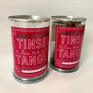 May include: Two clear plastic containers with silver lids and bases. Each container has a red label with the text "DON'T Get Your TINSEL in a TANGLE" in white and pink. The containers are for holiday survival.