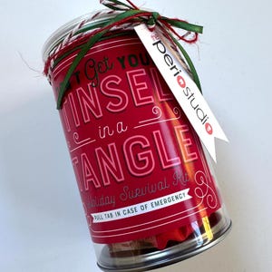 May include: A cylindrical container with a red label that reads "Don't Get You Tinsel in a Tangle" and "Holiday Survival Kit." The container is clear, revealing its contents, and is tied with green and red ribbon. A white tag reads "the periostudio."
