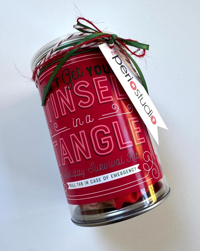 May include: A cylindrical container with a red label that reads "Don't Get You Tinsel in a Tangle" and "Holiday Survival Kit." The container is clear, revealing its contents, and is tied with green and red ribbon. A white tag reads "the periostudio."