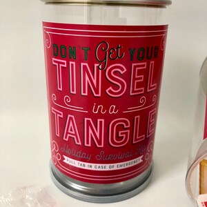May include: A clear plastic container with a red label that reads "DON'T Get YOUR TINSEL in a TANGLE". The container is a holiday survival kit, with a pull tab for emergencies. Candy cane and chocolate are visible.