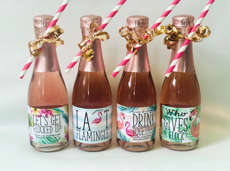 May include: Four mini champagne bottles with pink liquid, each adorned with a pink and white striped straw and gold ribbon. The labels feature tropical floral designs and phrases like "Let's Get Flocked Up!" and "LA Flamingle."