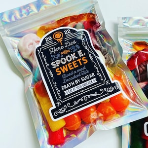 May include: Holographic cellophane bags filled with assorted Halloween sweets. Each bag features a label with the text "Here Lies Spook E. Sweets, Death by Sugar, Eat at Your Own Risk."