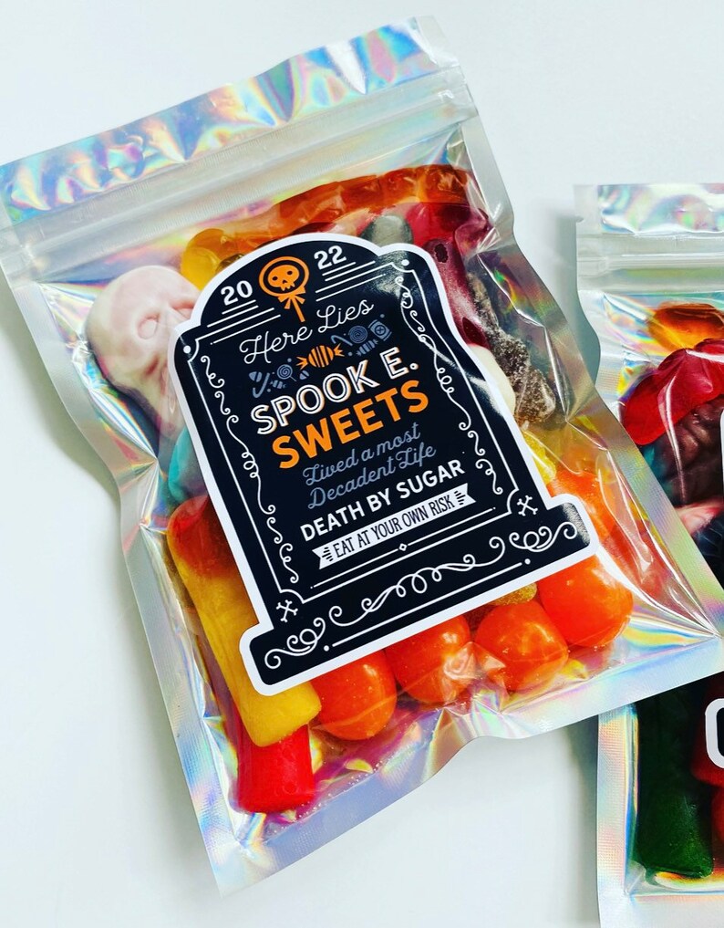 May include: Holographic cellophane bags filled with assorted Halloween sweets. Each bag features a label with the text "Here Lies Spook E. Sweets, Death by Sugar, Eat at Your Own Risk."