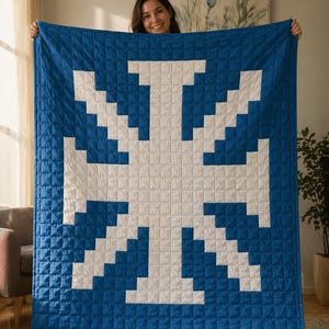 May include: A blue and white quilt with a geometric design. The quilt is a square shape and features a large white cross-like pattern against a blue background. The quilt is held up by a person.