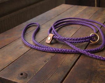 May include: A purple braided leash featuring a round gold-toned metal ring and a tan leather accent piece secured with a gold-toned metal rivet. The leash is constructed from a woven cord material and is coiled on a dark wooden surface.