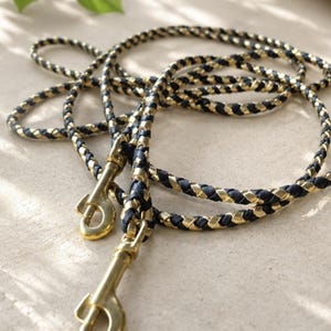 May include: A braided lead featuring alternating strands of dark navy blue and metallic gold material. The lead is equipped with two polished brass swivel snap hooks at the ends, designed for secure attachment, showcasing a durable and flexible construction.