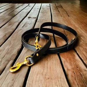 May include: A black leather dog lead with gold-coloured metal clasps and fittings. The lead is coiled on a wooden surface, highlighting its sleek design and quality craftsmanship. The fittings include a swivel clasp for easy attachment to a dog's collar.