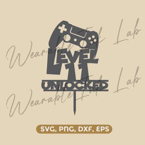 Pode incluir: Um topo de bolo cinza-escuro com a silhueta de um controle de videogame acima do texto LEVEL 11 UNLOCKED. O design inclui uma haste pontiaguda na parte inferior para inserção. A imagem inclui o texto SVG, PNG, DXF, EPS na parte inferior.