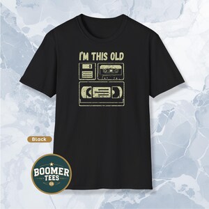 May include: Black t-shirt featuring the phrase "I'M THIS OLD" and images of a floppy disk, cassette tape, and VHS tape. The shirt has a crew neck and short sleeves. The Boomer Tees Co. logo is at the bottom.