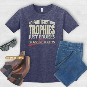 May include: Heather navy t-shirt with the text "NO PARTICIPATION TROPHIES JUST BRUISES AND BRAGGING RIGHTS". The image also includes a pair of sunglasses, brown boots, and blue jeans.