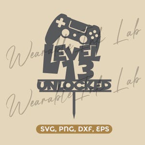 Pode incluir: Um topo de bolo cinza-escuro apresentando a silhueta de um controle de videogame acima do texto LEVEL 13 UNLOCKED. O design inclui uma haste vertical na parte inferior. A imagem também exibe o texto SVG, PNG, DXF, EPS na base.