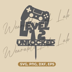 Pode incluir: Um topo de bolo cinza-escuro com a silhueta de um controle de videogame acima do texto LEVEL 12 UNLOCKED. O design inclui uma haste pontiaguda na base para inserção. Abaixo do design, uma etiqueta retangular exibe SVG, PNG, DXF, EPS.