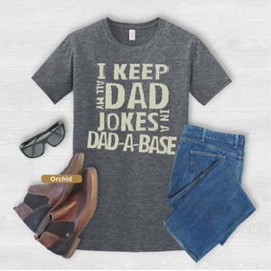 May include: Grey t-shirt with the text "I KEEP ALL MY DAD JOKES IN A DAD-A-BASE". The image also includes blue jeans, brown leather boots, and sunglasses.