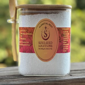 Puede incluir: Un tarro de cristal lleno de sales de baño blancas, con tapa y cuchara de madera. El tarro tiene una etiqueta que dice "Soulseed Solutions" y "Celestial Rest". La etiqueta también enumera los ingredientes y las instrucciones de uso.