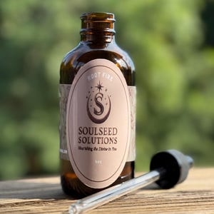 Puede incluir: Una botella de vidrio marrón con una etiqueta que dice "ROOT FIRE SOULSEED SOLUTIONS Nourishing the Divine in You" y una capacidad de 118 ml. Un cuentagotas de vidrio transparente con tapa negra descansa junto a la botella. El fondo es verde borroso.