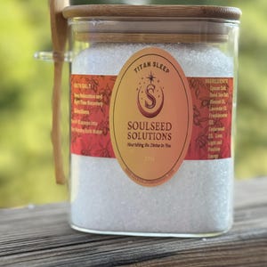 Puede incluir: Un tarro de vidrio transparente lleno de sales de baño blancas, con una tapa de madera y una cuchara de madera. El tarro tiene una etiqueta con las palabras "Titan Sleep" y "Soulseed Solutions". La etiqueta enumera ingredientes.
