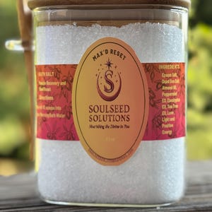 Puede incluir: Un tarro de vidrio transparente lleno de sales de baño blancas, cubierto con una tapa de madera. El tarro tiene una etiqueta con el texto "SOULSEED SOLUTIONS" y "MAX'D RESET". Una cuchara de madera descansa junto al tarro.