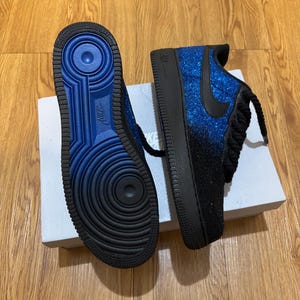 May include: Pair of blue and black Nike Air Force 1 sneakers. The sneakers have a glittery blue upper that fades to black, a black swoosh, and black laces. The soles are black with a blue interior.
