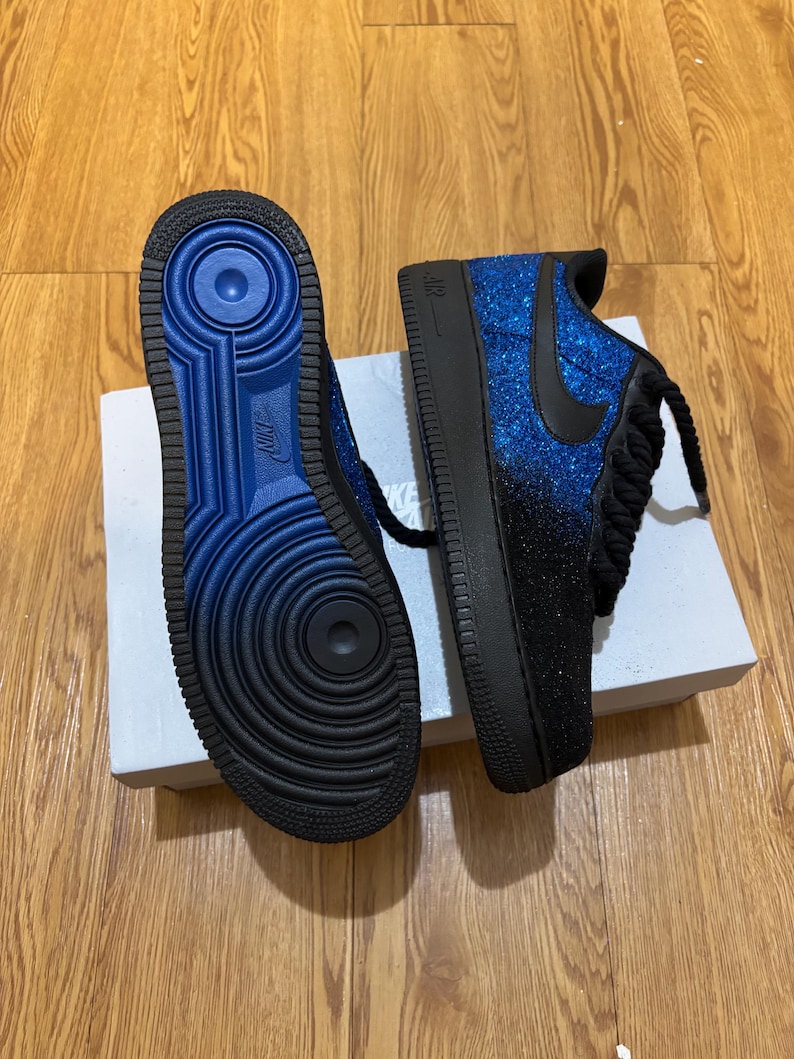 May include: Pair of blue and black Nike Air Force 1 sneakers. The sneakers have a glittery blue upper that fades to black, a black swoosh, and black laces. The soles are black with a blue interior.