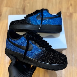 May include: A pair of black and blue glitter sneakers. The sneakers have a black sole, laces, and Nike swoosh. The upper transitions from black to blue glitter. The shoes are displayed on a white box.