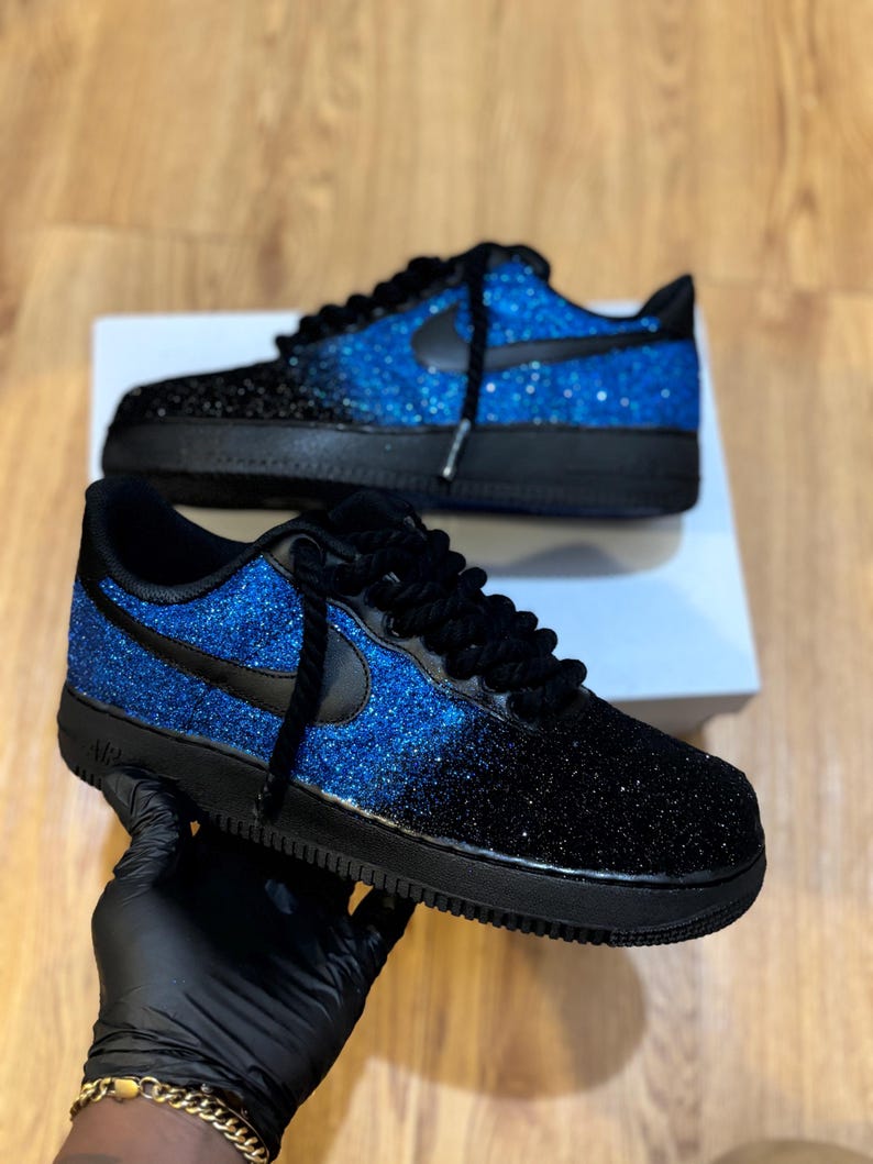 May include: A pair of black and blue glitter sneakers. The sneakers have a black sole, laces, and Nike swoosh. The upper transitions from black to blue glitter. The shoes are displayed on a white box.