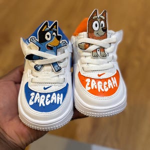 Puede incluir: Un par de zapatillas blancas y azules personalizadas con parches de personajes de perros de dibujos animados. El zapato azul tiene una puntera azul con el nombre "ZAREAH" en blanco. El zapato naranja tiene una puntera naranja con el nombre "ZAREAH" en blanco.