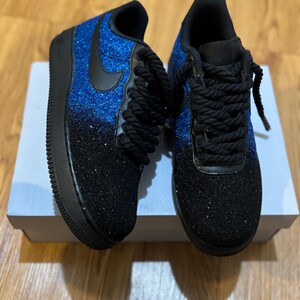 May include: A pair of custom sneakers with a blue and black glitter design. The shoes have black laces, a black swoosh, and a black sole. The glitter fades from blue at the top to black at the bottom. The shoes are sitting on a white box.