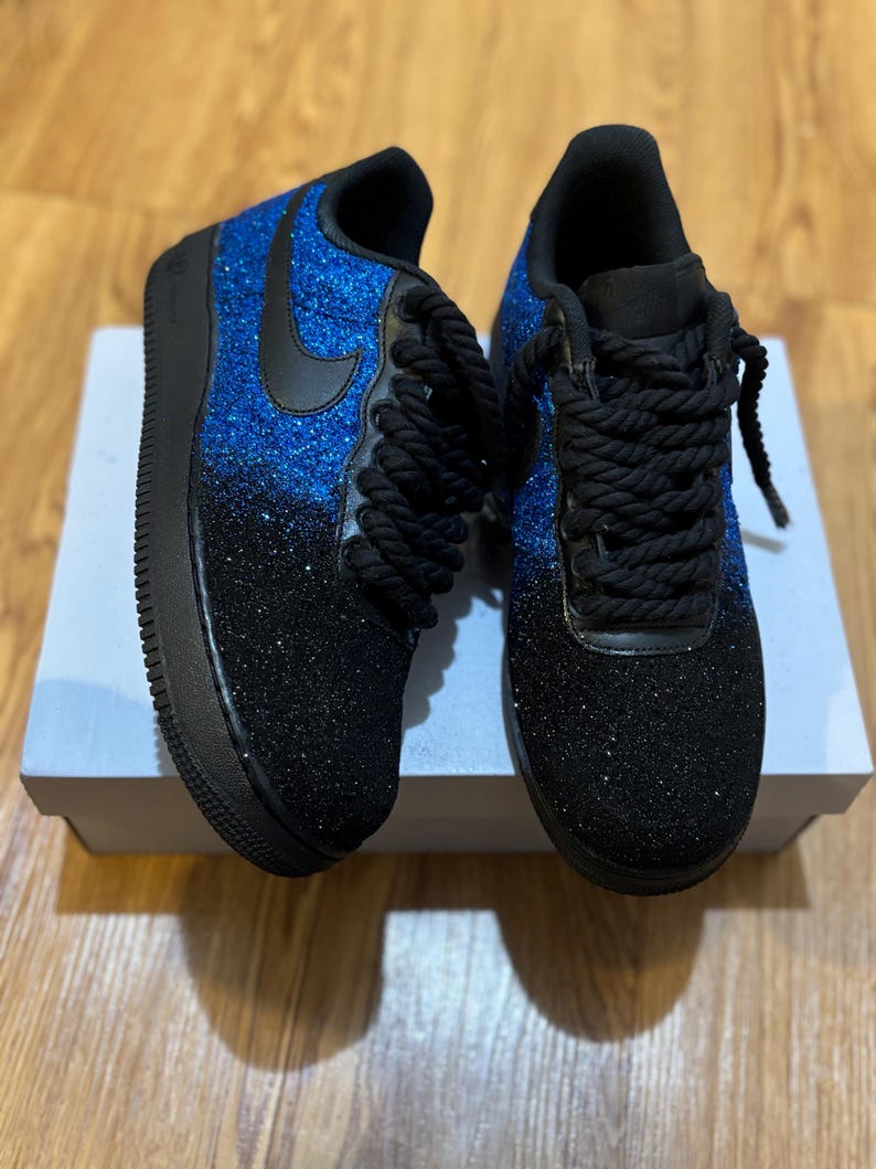 May include: A pair of custom sneakers with a blue and black glitter design. The shoes have black laces, a black swoosh, and a black sole. The glitter fades from blue at the top to black at the bottom. The shoes are sitting on a white box.