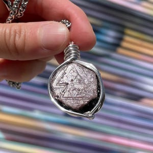May include: A silver-toned pendant necklace featuring a faceted, light purple gemstone. The stone is encased in a silver wire wrap and hangs from a delicate chain. The gemstone has a geometric pattern on its surface.