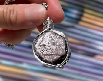May include: A silver-toned pendant necklace featuring a faceted, light purple gemstone. The stone is encased in a silver wire wrap and hangs from a delicate chain. The gemstone has a geometric pattern on its surface.